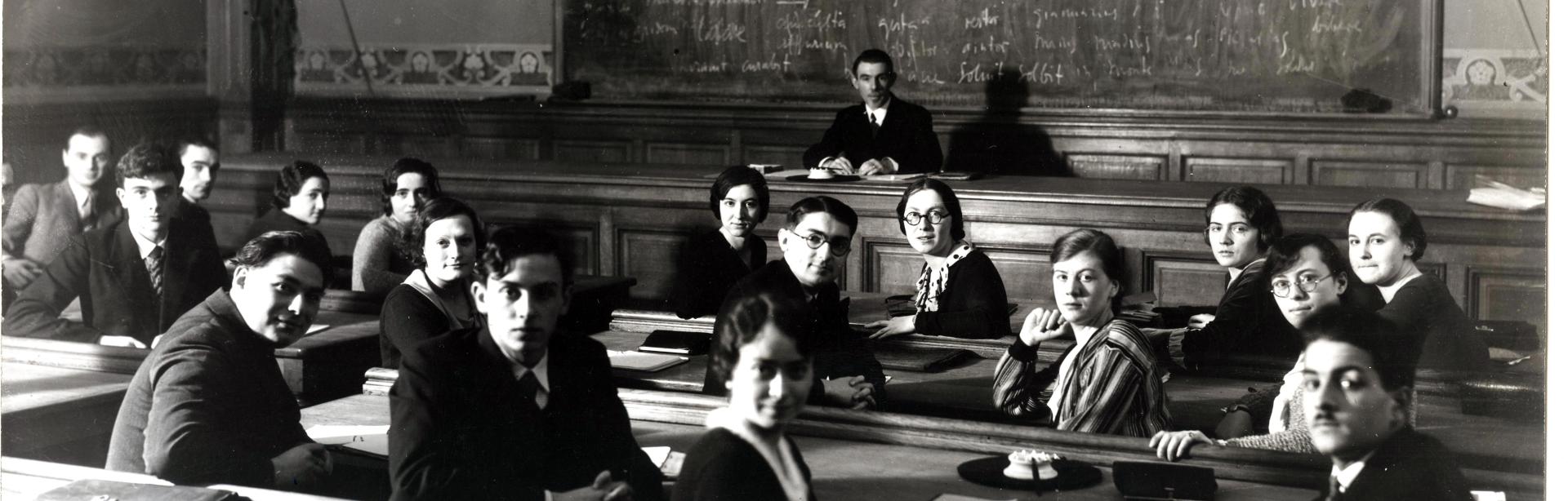 Des élèves assis en classe se retournent pour poser, le professeur Clovis Brunel est assis au fond à la chaire
